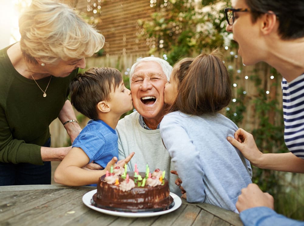 Senior aged male celebrates birthday outdoors with his family, kisses on his cheek from young children, and a birthday cake