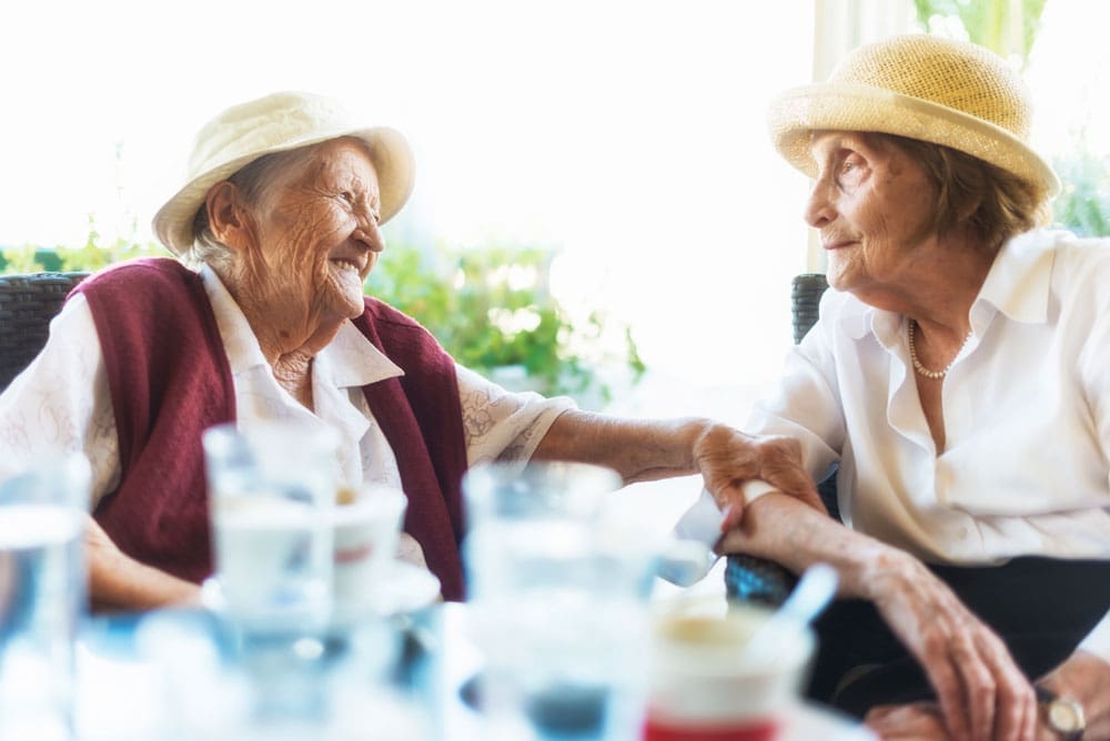 Two female senior living residents in hats sit at outdoor table and smile at each other 